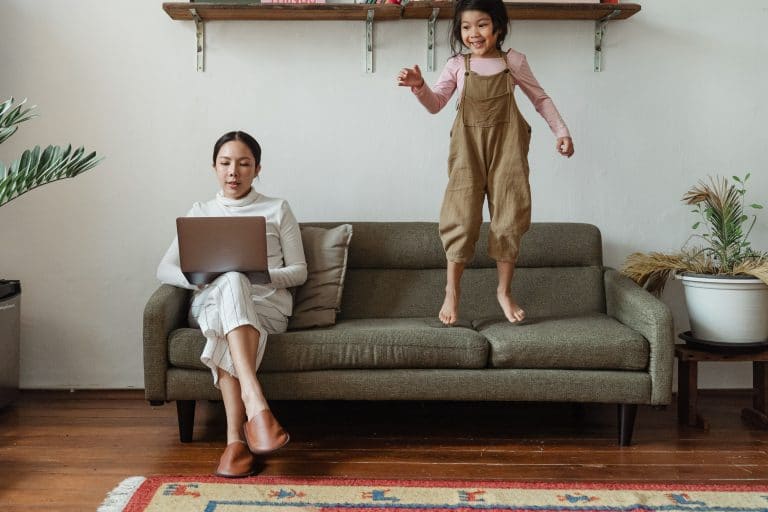 Illustration d'une femme travaillant sur son ordinateur avec sa fille sautant de joie sur le canapé, symbolisant la recherche d'équilibre entre travail et vie personnelle.