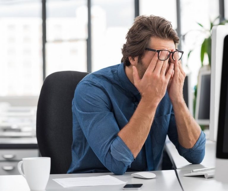 Un homme au bureau, portant des lunettes, se frotte les yeux tout en relevant légèrement ses lunettes. Son expression montre des signes de stress et de fatigue au travail.