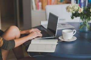 Une femme concentrée à sa table de travail avec un ordinateur, un café et un carnet en train de se former en continue.